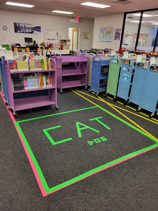 book carts full of books waiting to be sent out to Library locations stand in a row. In front of them, brightly colored tape on the ground inexplicably spells out the word "cat" in huge letters, and "dog" in smaller letters.