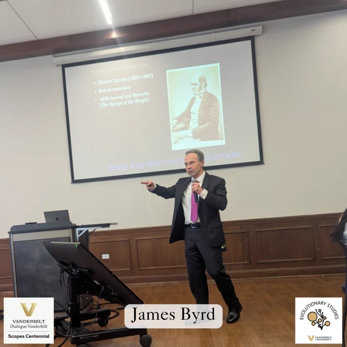 James Byrd, dressed in a dark suit with a purple tie, gestures while giving a presentation on Charles Darwin and evolution at the Scopes Centennial event at Vanderbilt University. A slide behind him displays an image of Charles Darwin with bullet points summarizing his contributions, including his 1839 journal and remarks from The Voyage of the Beagle.