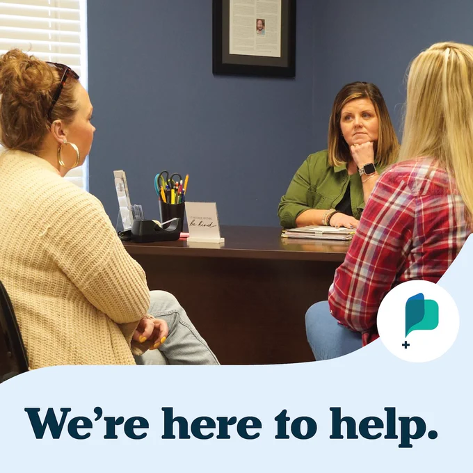 Three people in a discussion at a desk in an office setting, with a sign that reads "We're here to help.