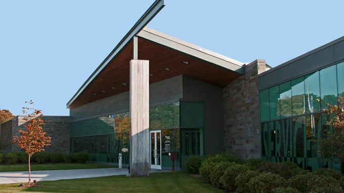 Exterior view of a modern Hardwood Creek Library building with large glass windows and a unique wooden and stone facade, featuring a prominent overhang roof and a small tree in the foreground.