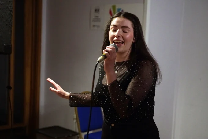 A woman singing into a microphone on stage, engaging the audience at The Beacon open mic night.