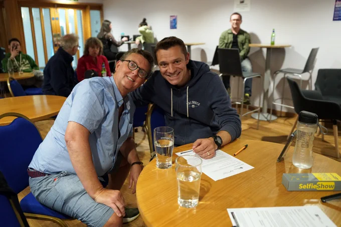 A comedy duo smiling for the camera, with a lively crowd visible in the background at The Beacon Open Mic.