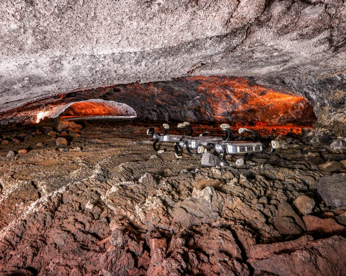A photograph taken underground in a 'lava tube' which is a tunnel created long ago by flowing lava. Inside the rocky tunnel is a robotic rover explorer that's traversing the harsh terrain. 