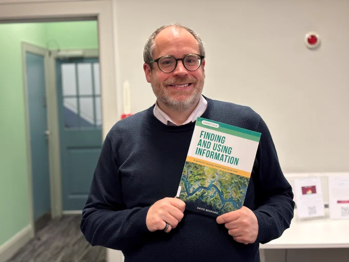 A man smiles while holding a copy of the book 'Finding and using information'.