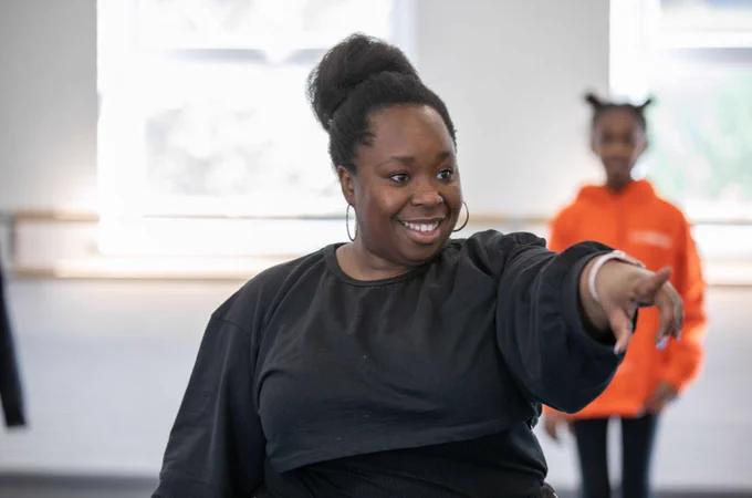 Photo of Eleanor Manners, incoming Artistic Director of Theatre Centre. Eleanor is in a rehearsal room, smiling as she points towards something across the room, behind the camera. A young person stood in the background behind her. Photo credit: Oliver Dixon.