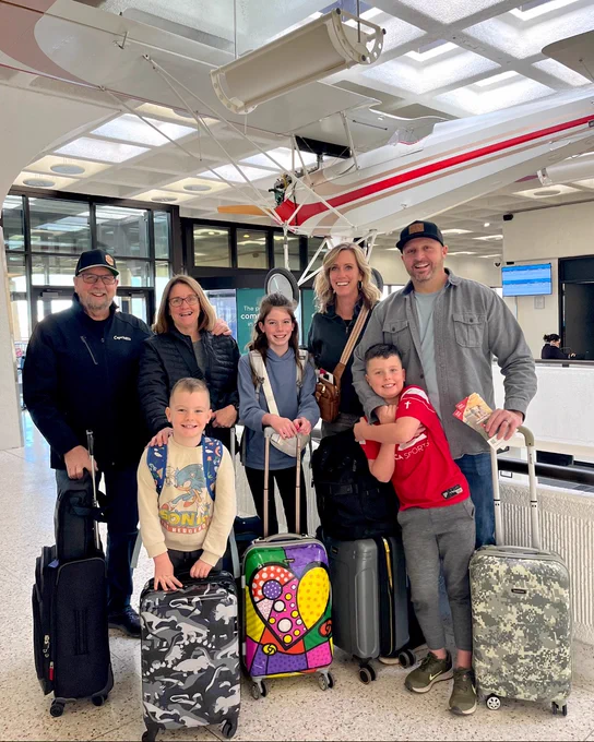 Group of seven individuals with luggage at an airport terminal, smiling and posing for a photo.