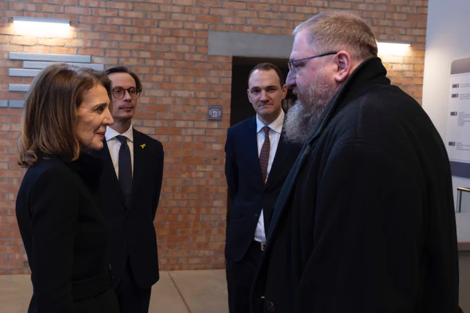 Four people (a woman and man in the foreground, and two man in the background) are standing inside a space with a red brick wall