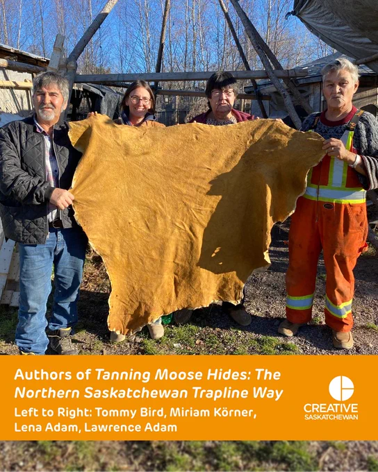 Two men and two women holding up a tanned moose hide on a sunny day. An orange bar across the bottom reads “Authors of Tanning Moose Hides: The Northern Saskatchewan Trapline Way Left to Right: Tommy Bird, Miriam Körner, Lena Adam, Lawrence Adam” 
