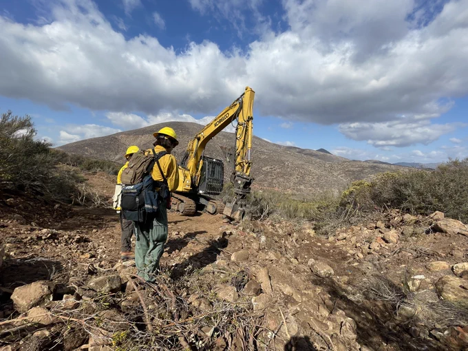 An excavator  works in a dozer-created fire break in a mountainous area. Two resource advisors in protective clothing are nearby. 