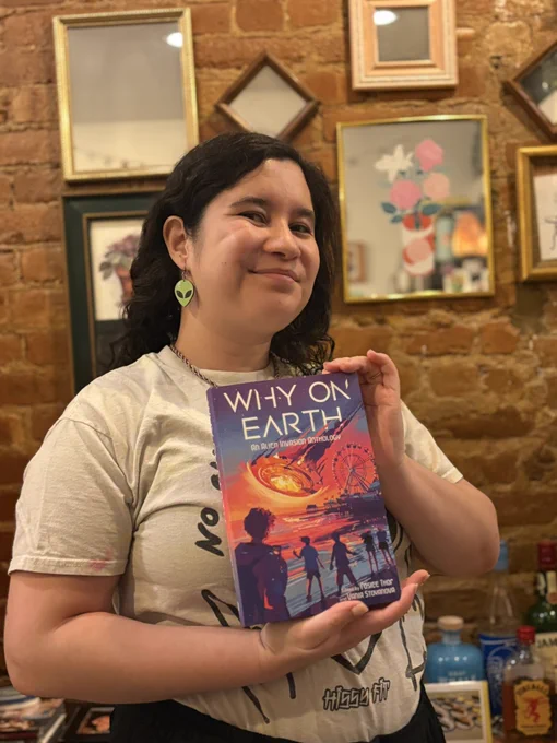 me smiling with a why on earth final copy, wearing a shirt that features an alien flipping the middle finger that says “no one is illegal,” and an alien earring