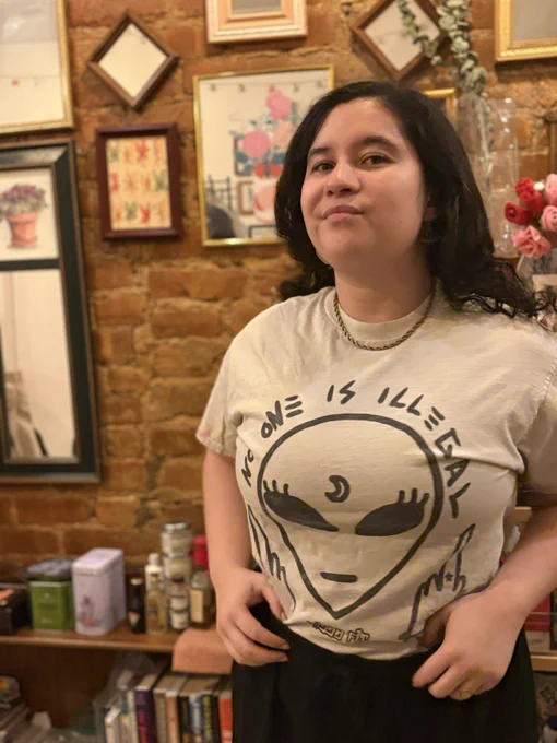 me with a small smile looking at the camera. my shirt says “no one is illegal” over an alien giving two middle fingers.