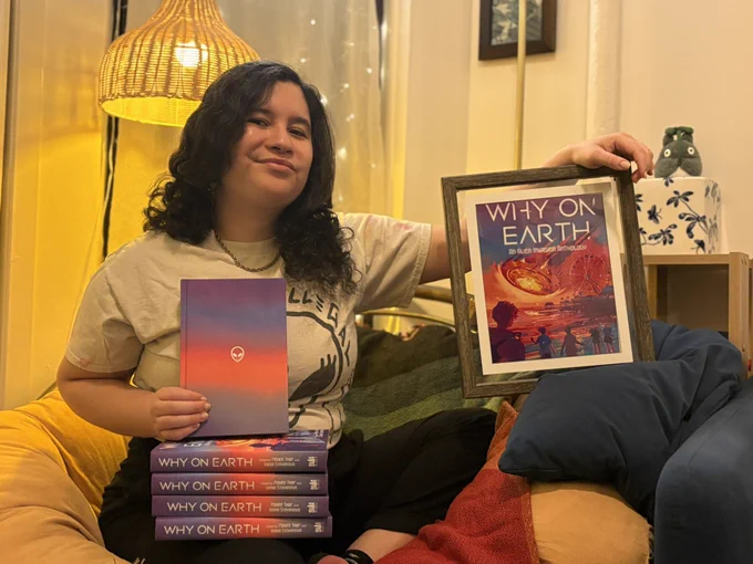 me smiling with a stack of why on earth final copies, wearing a shirt that features an alien flipping the middle finger that says “no one is illegal,” and an alien earring, holding up a poster of the jacket set and a beautiful hardcover without its jacket, showing off its blue, purple, orange pink colors and an alien head