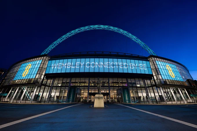 Wembley Stadium, with the EE logo either side of the famous arch lit up in aqua. "Wembley Connected by EE" is writ large on the LED screen. 