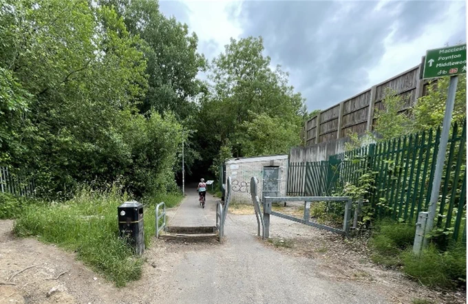 Photograph shows a paved traffic-free route with three sets of barriers across it: (1) a horsestyle consisting of two beams about 20 cm high, a K frame which narrows the width of access, and a metal gate. To the edge is a desire line going round the barriers, but the surface is uneven, meaning it can be unsuitable for some people using mobility aids.
