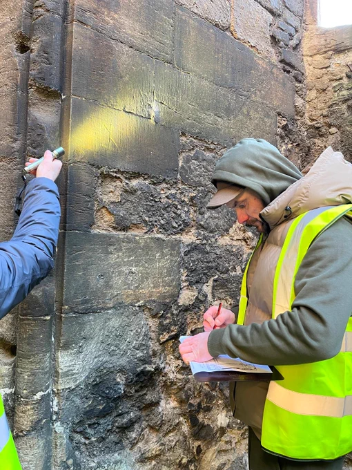A person shines a torch on a mason's mark, and another person in high vis makes a drawing of it.