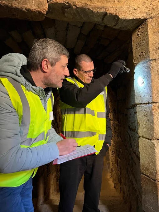 A person shines a torch on a mason's mark, and another person in high vis makes a drawing of it.