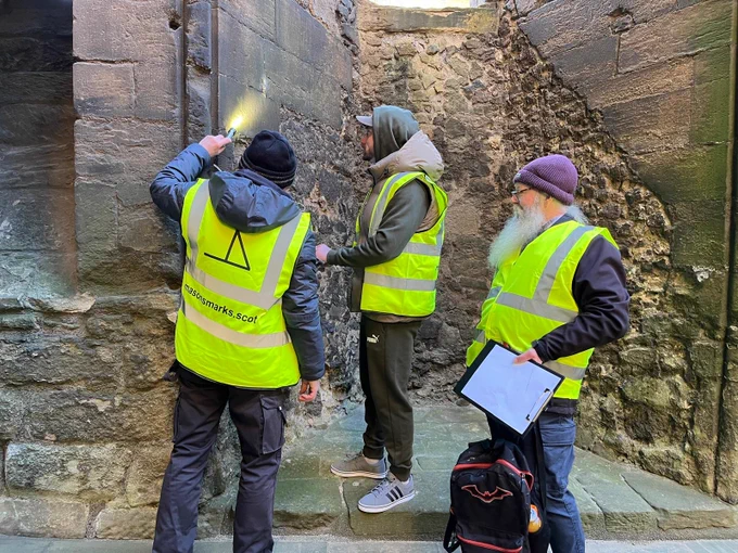 A person in high via shines a torch on a mason's mark, and another two people in high vis holding clipboards make drawings of it.