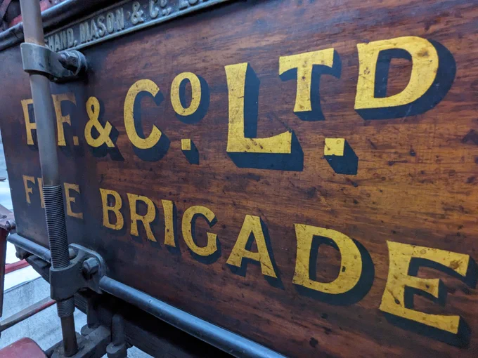 Close up of the wooden central section of a piece of fire brigade equipment made by Shand Mason. Handwritten letters have been painted onto the side of the wooden section in yellow, with black shadowing. This spells out P. F. & Co. Ltd. Fire Brigade