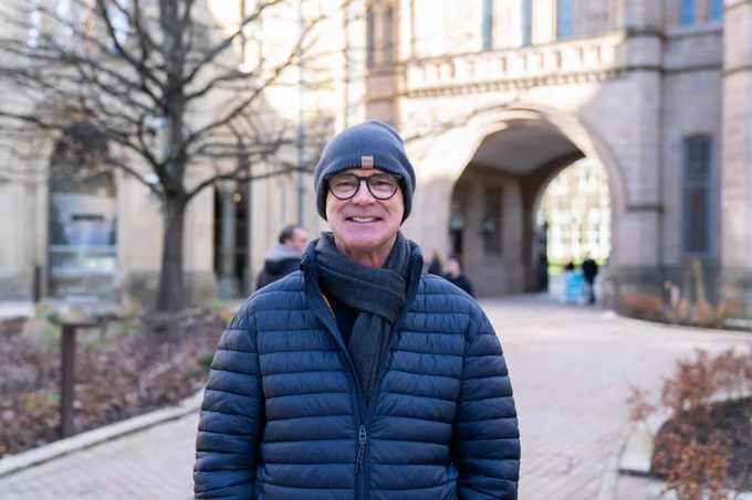 Professor Jamie Woodward, wearing a dark blue coat, jeans and beanie hat, stands in the Old Quad on the University of Manchester campus with his hands in his pocket
