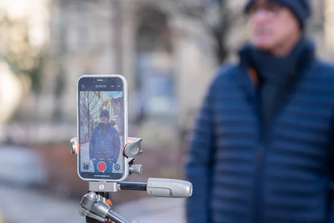 Professor Jamie Woodward, wearing a dark blue coat, jeans and beanie hat, stands in the Old Quad on the University of Manchester campus being filmed. A phone is recording a video, on a tripod.