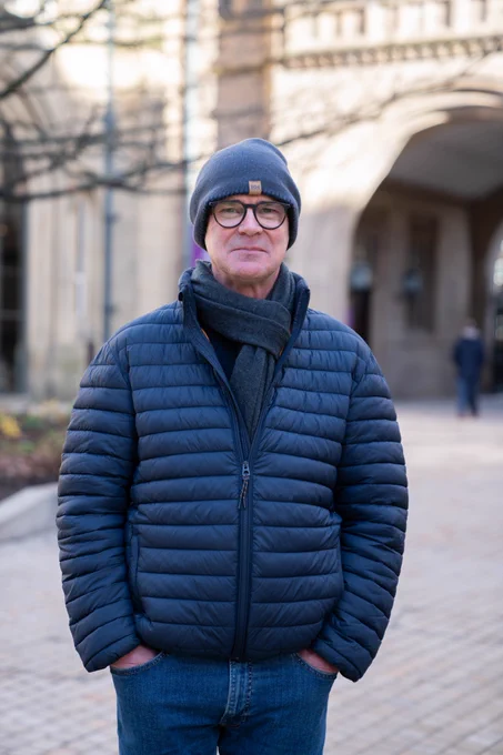 Professor Jamie Woodward, wearing a dark blue coat, jeans and beanie hat, stands in the Old Quad on the University of Manchester campus with his hands in his pocket