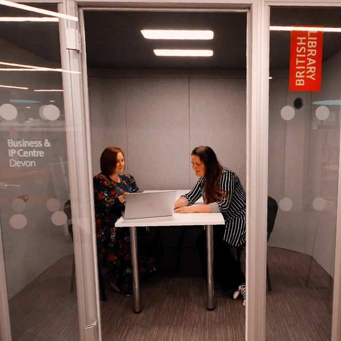 Two people sat opposite sides of a table, sharing a laptop.  They are in a bookable 'pod'. 