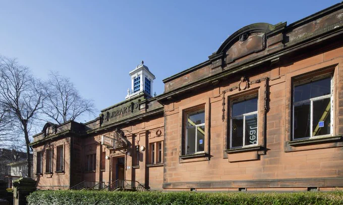 Exterior shot of Langside Library, a red sandstone single-storey Victorian building with white tower on the roof. The background is trees on the left-hand side and blue sky.