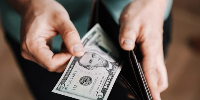 A person taking a five dollar bill from a wallet symbolizing personal finance and budget management.