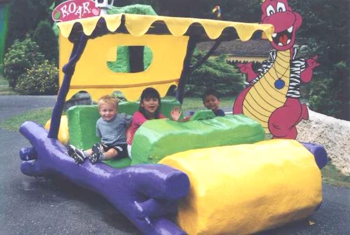 This photo depicts a fun and colorful scene at what appears to be a themed amusement park or attraction. In the foreground, there is a whimsical vehicle designed to look like a stone-age car from the "Flintstones" cartoon, with large, round wheels and a structure made to resemble logs and rocks. The car is painted in vibrant colors, primarily yellow, green, and purple, with the word "ROAR" displayed on a sign at the front.

Three children are seated in the car, looking excited and happy. They are dressed casually, suggesting a family-friendly environment. Behind the car, there is a large, cartoonish dinosaur figure, likely a statue or a ride mascot, adding to the prehistoric theme. The dinosaur is pink with a yellow belly, wearing a zebra-striped vest, and has a friendly, animated expression.