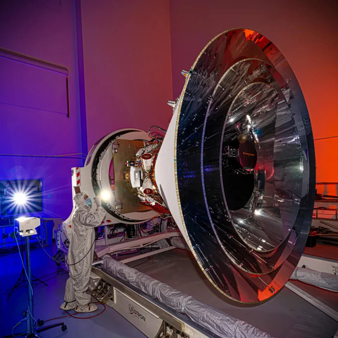 NASA’s SPHEREx observatory undergoing integration and testing at BAE Systems in Boulder, Colorado, in April 2024. The image shows the observatory with a polished and reflective surface in a brightly lit testing facility, with an American flag in the background, surrounded by technical equipment and a vibrant blue and red glow from the lighting.
