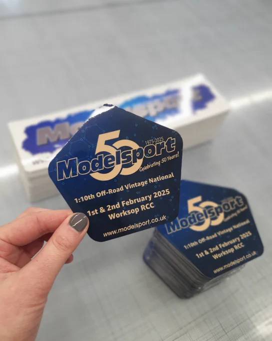 A pile of Modelsport logo bumper stickers and pentagon shaped 50th anniversary stickers on a workbench.  A hand in the foreground holding one of the 50th anniversary stickers.