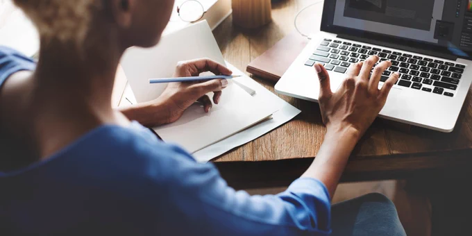 A woman sits in front of a computer while taking notes in a notebook.