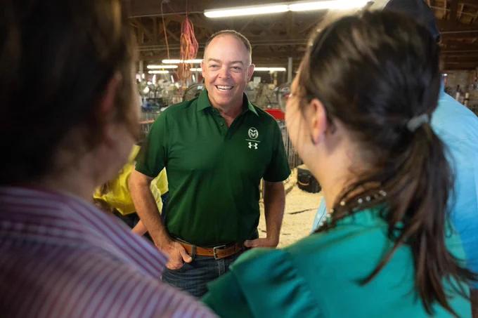 Don Thorn, CSU's manager of external relations for rural Colorado, smiles while talking to two people at the Colorado State Fair. Don is wearing a green collared shirt with the CSU logo on it, a brown belt and blue jeans, while the two people he's interacting with are wearing a striped shirt and a teal shirt; their faces are turned from the camera.