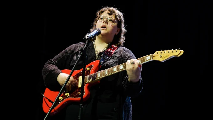 A young person plays the guitar onstage, singing into a microphone. The guitar is red. The young person wears black clothing, a cardigan and jeans, with a black shirt. They have glasses on.