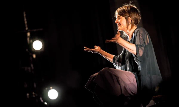 A woman sit on stage with her arms open towards the audience.
