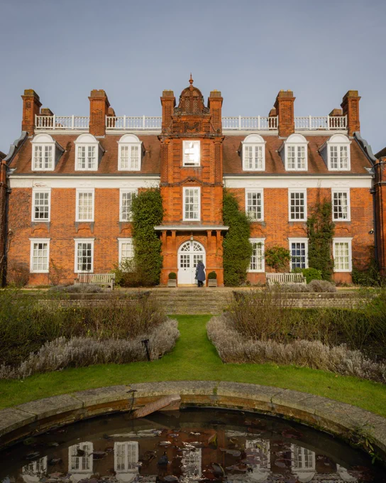A three storey red brick building with white windows, in front of a large pond with a reflection.