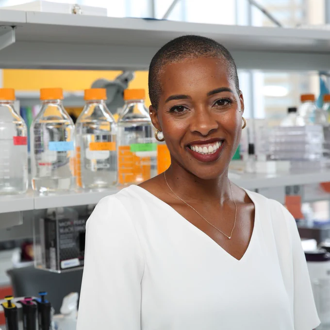 Dr. Marlin, a Black woman neuroscientist, is smiling proudly in her lab space. She’s wearing a white shirt, simple necklace, small hoop earrings, dark red lipstick and has a beautifully shaved head.