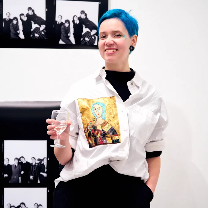 An image of someone enjoying  the launch night for For Your Pleasure: 15 Years of DuoVision at the Open Eye Gallery. They are stood, with one hand in their pocket, one hand holding a glass, smiling. They have a white shirt on, with a sketch of somebody on one side of the shirt. They have sparkley eye make up on! In the background, there is some images, blurry, presumably part of the exhibition. 