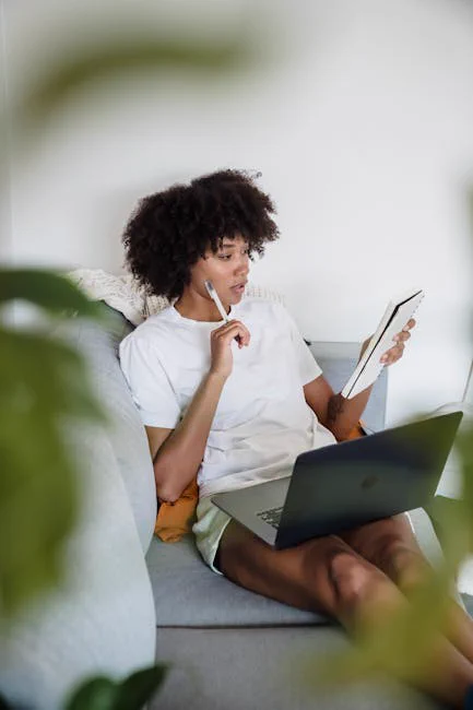 Focused young woman studying at home with a laptop and notepad, sitting comfortably on a couch.