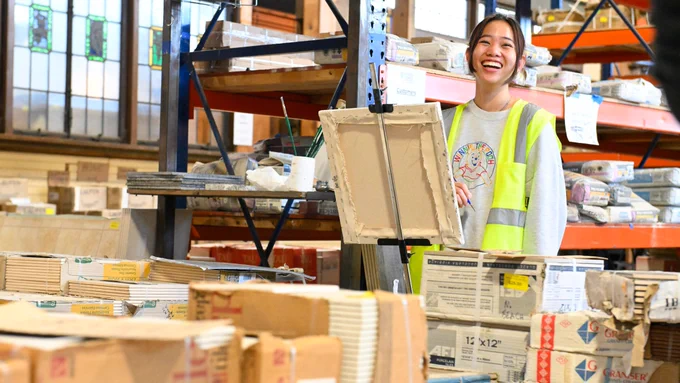 A smiling worker wearing a safety vest carries a cardboard box in a well-stocked warehouse.