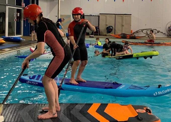 Two people stand on paddleboards in a swimming pool. There are children and adults behind playing on paddleboards too.