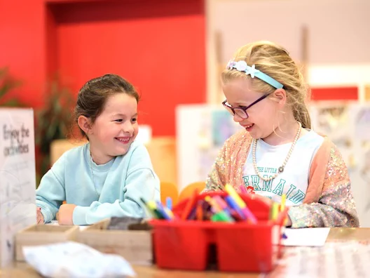 A photograph of two children smiling and engaging with each other at a table, with a basket of colourful supplies between them.