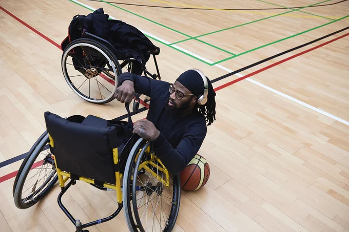 A person is making adjustments to a wheelchair on a basketball court, with a basketball next to them. The wheelchair features yellow accents and the court has multiple colored lines.