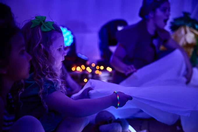 Image description: A young girl is in a creative space with a couple of additional participants. She is holding one edge of large piece of fabric and a woman in the background is holding another section of the fabric.