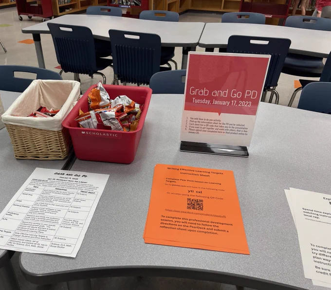 A tabletop with some bins of snacks, a "Grab and Go PD" sign and some stacks of paper with instructions and QR codes.