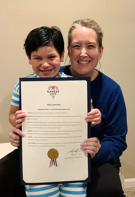 Leah crouches down next to her son Caden. Both are smiling and holding a printed proclamation declaring Feb. 15 as International Angelman Day in Kansas City.