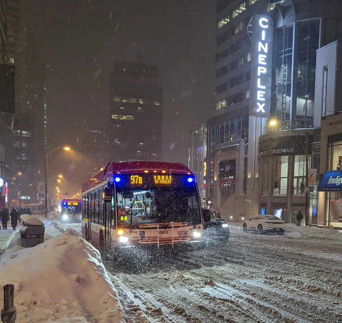 two buses in a row on a snowy city street, as snow falls during the night