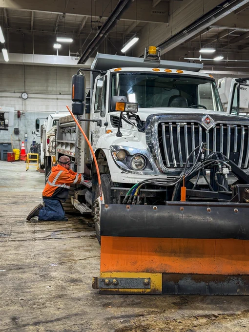 A mechanic kneels on the floor next to a vehicle in a large garage, surrounded by heavy machinery trucks. 