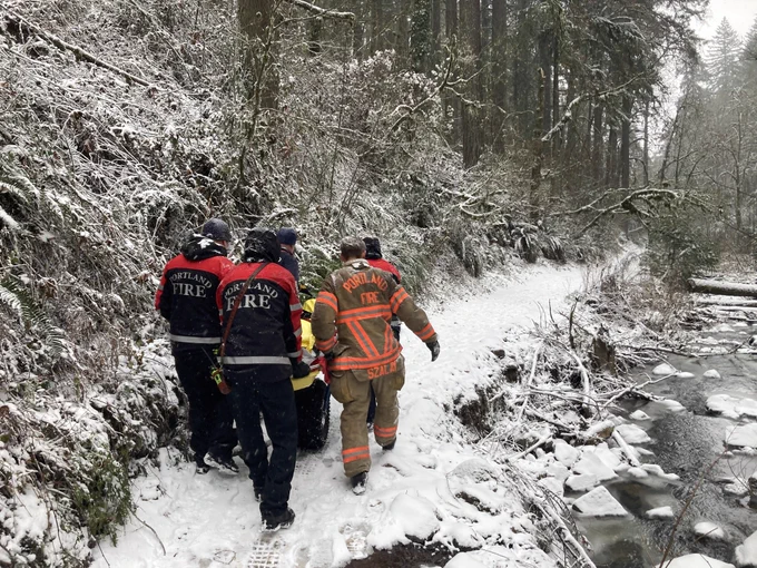 Firefighters carry an injured hiker on a snow-covered, forested trail. 
