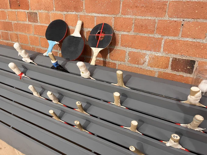 Slatted grey rack against a brick wall holds a range of adapted table tennis bats with handles protruding. On the top edge, three adapted bats stand upright against the wall - one has a moulded handle with an extension; one with two cut out finger  grooves on the handle and central hole in the bat with a crossed tape to catch the ball; third with a widened handle